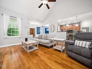 A living room with hardwood floors, a light gray sofa and matching loveseat, a wooden coffee table and side tables, a reclining chair, white drapes on a window, and an open view into a kitchen with brown cabinets.