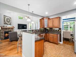 Open kitchen with wooden cabinets, black countertops, and tile flooring. Adjacent living area features a stone fireplace, TV, armchair, and visible dining room with windows in the background.