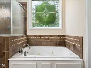 A white built-in bathtub with silver faucets is set in a tiled surround. Above the tub is a decorative window with frosted and stained glass. Part of a glass shower enclosure is visible to the left.