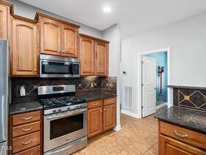 A kitchen with wooden cabinets, stainless steel gas stove and microwave, black tile backsplash with tan accents, granite countertops, tiled floor, and an open door revealing a utility room.