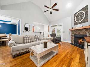 A living room with a light beige sofa, wooden coffee table, armchair, stone fireplace with a clock above it, hardwood floors, and a ceiling fan. There is an open doorway to a dining area and kitchen.