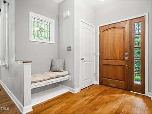 A wooden front door with glass panels is next to a small alcove with a built-in bench, cushion, and pillow. The floor is hardwood, and a white door and decorative stained glass window are visible.