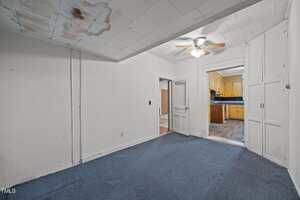 A room with blue carpet, white walls, a stained ceiling, built-in cabinets, and a ceiling fan, leading to a hallway and a kitchen with yellow cabinets and dark countertops.