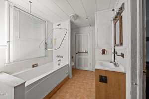 A bathroom at 107 Franklin Street, Oxford features a white bathtub and shower, a small white sink with a mirror, white walls and ceiling, wood-patterned vinyl flooring, a window with blinds, and a closed white door in the background.