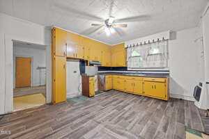 A kitchen with yellow cabinets, a dark countertop, wood-style flooring, a ceiling fan with a light, and two small windows with white curtains above the sink. The space appears worn and somewhat outdated.