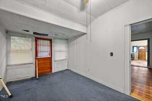 A small, empty room with white walls, a blue carpet, and a wooden door with two windows. The ceiling is slanted with visible water damage, and there is an open doorway leading to another room with hardwood flooring.