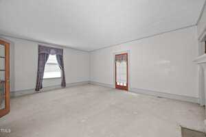 Empty room with light-colored carpet, white walls, and a white ceiling. There is a window with purple curtains, a wooden door with a matching curtain, a glass-paned door, and a fireplace on the right side.