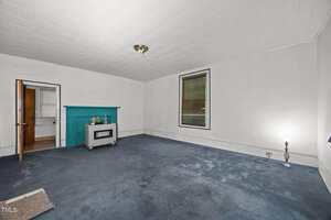 A mostly empty room with white walls, blue carpet, a single window, a turquoise fireplace, a small heater, an open door to another room, and a floor lamp in the corner.