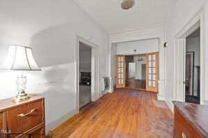 A hallway with hardwood floors, a table with a lamp on the left, white walls, and multiple doorways, including double wooden doors with glass panels leading to a room in the background.