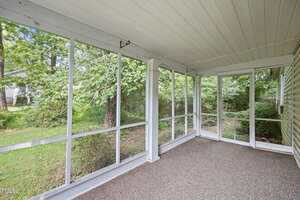 A screened-in porch with white framing and a carpeted floor, overlooking a green backyard with trees and grass. The porch has large screened windows and a white ceiling.