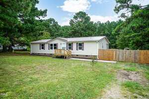 A single-story manufactured home with cream siding and purple shutters, surrounded by trees. The house has a small wooden porch, a fenced backyard, and sits on a grassy yard under a partly cloudy sky.