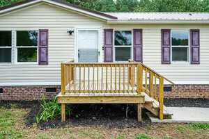 A beige house with purple shutters features a small wooden porch with steps and a railing leading to a white door. The porch is built over a mulched area with some plants. Brick foundation is visible below the siding.