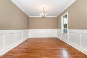 An empty room with light brown walls above white wainscoting, hardwood floors, a white ceiling, a window on the right wall, and a ceiling light fixture with five bulbs in the center.