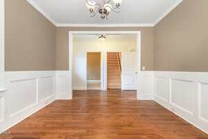 A room with hardwood floors, beige walls with white wainscoting, and a chandelier. An open doorway leads to a hallway with carpeted stairs and two closed doors. The space is empty and well-lit.