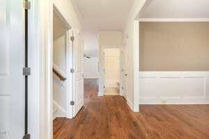 A hallway with wooden flooring leads to a bathroom with white fixtures. Two open doors are visible on the left; one reveals stairs with a beige carpet. The right side features white wainscoting and a beige wall.