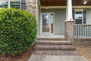 A front porch with brick steps, a brick and stone exterior, a large shrub on the left, white columns, and a wooden ceiling. The front door has a glass storm door, and there are two windows with black shutters.