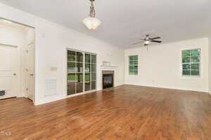 Spacious living room with hardwood floors, white walls, a ceiling fan, a fireplace, and large windows. Glass sliding doors lead to a covered porch. Light fixture hangs from the ceiling. Doorway visible on the left.