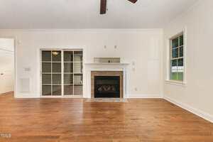 A living room with hardwood floors, a white fireplace with a stone surround, a glass sliding door to the left, a single window to the right, and cream-colored walls with white trim and crown molding.
