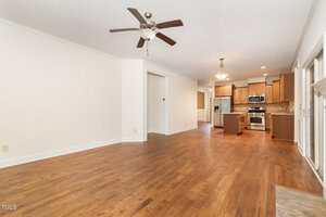 Open-concept living area with hardwood floors, a ceiling fan, neutral walls, and a view into a modern kitchen with wooden cabinets, stainless steel appliances, and recessed lighting. Large windows line the right side.