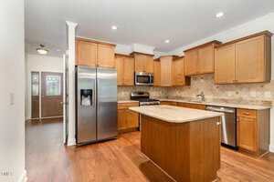 Modern kitchen with stainless steel appliances, light wood cabinets, granite countertops, a center island, and hardwood floors. There is a double door refrigerator, gas stove, microwave, and a dishwasher. Entryway is visible in the background.