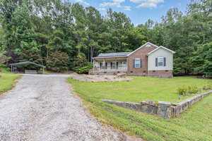 A single-story brick house at 824 Kittrell Road features a covered front porch, nestled among tall trees. A gravel driveway leads to a carport on the left, with a small stone retaining wall in the front yard.