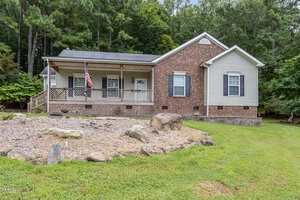 A single-story house with a front porch, brick and siding exterior, an American flag, and a bare flower bed with rocks in the yard. There are trees and greenery in the background.