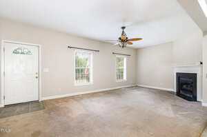 A mostly empty living room with beige carpet, two windows, a white front door on the left, a fireplace on the right, a ceiling fan with lights, and light tan walls. Some stains and wear are visible on the carpet.