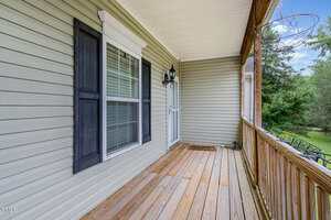 A wooden porch with a light brown floor, beige siding, a window with black shutters, a white storm door, a doormat, an ornate metal light fixture, and a wooden railing overlooking green trees.