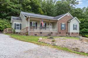 Single-story house with a brick and siding exterior, a covered front porch displaying an American flag, and a gravel driveway, surrounded by grass, trees, and a mulched garden area.