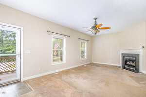 A mostly empty living room with beige walls, carpeted floor, two windows with blinds, a ceiling fan, a fireplace in the corner, and a door leading to a wooden deck outside.