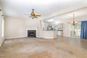 Open living area with beige carpet, ceiling fan, and fireplace in the foreground; adjacent kitchen with white appliances and bar counter; dining area with chandelier and sliding glass door to outside.
