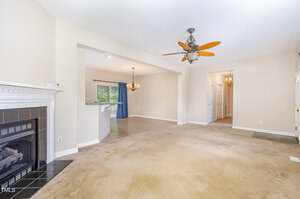 A living room with beige carpet, a ceiling fan with lights, fireplace with black tile surround, and an open view into a dining area with tile floor, chandelier, and sliding glass door with blue curtains.