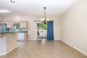 An empty dining area with a tile floor and chandelier, next to a kitchen with light wood cabinets and white appliances. A sliding glass door with a blue curtain opens to a view of an outdoor patio and trees.