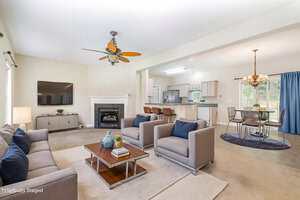 Open-concept living and dining area with neutral walls, modern furniture, a wall-mounted TV, fireplace, kitchen with bar stools, and a dining table under a chandelier near large windows with blue curtains.