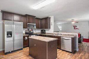 A modern kitchen with dark wood cabinets, stainless steel appliances, a center island, and marble-patterned countertops. The kitchen opens to a living area with a ceiling fan and a red and white rug.