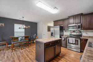 A modern kitchen with dark wood cabinets, stainless steel appliances, a center island, and a dining area with a wooden table and bench seating. The floor is wood, and the walls are painted gray and white.