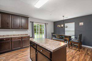 A kitchen with dark wood cabinets, a central island with a marbled countertop, and a dining area with an L-shaped bench and table. Large windows and glass doors provide natural light. The floor is wood-patterned.