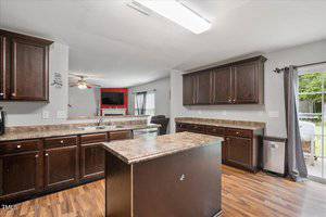 Spacious kitchen with dark wood cabinets, a central island, and marbled countertops. Stainless steel appliances and a sliding glass door to the outside are visible. The open layout connects to a living area with a TV and fireplace.