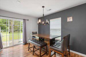A dining area with a dark wood table, two padded benches, and a chair on wood flooring. Large sliding glass doors and a window let in natural light; a modern chandelier hangs above the table.