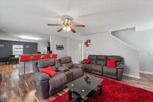 A living room with two brown reclining sofas, a black glass coffee table on a red rug, and a ceiling fan with lights. The space opens to a kitchen with red bar stools and wood flooring throughout. Stairs are visible to the right.