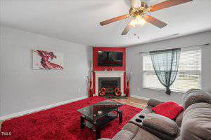A living room with gray walls, a red accent wall around a fireplace, a TV mounted above the fireplace, a red area rug, gray sofa, glass coffee table, and a ceiling fan with lights.
