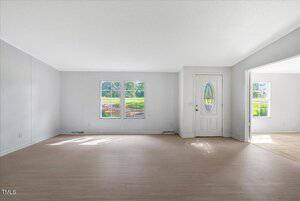 Unfurnished living room with light wood floors, white walls, two windows, and a white front door. Adjacent room with tile flooring and additional windows visible on the right.