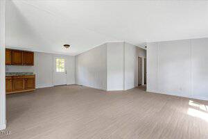 Unfurnished room with light-colored wood flooring, white walls, and a partially visible kitchen area with wooden cabinets. There is a door with a window, hallway, and natural light coming from the right side.