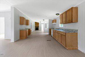A spacious, empty kitchen with light wood cabinets, marble countertops, and light-colored flooring. The room is well-lit by natural light from windows and open entryways, with a partial view of an adjacent room.