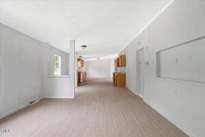 Unfurnished open-plan interior with light wood flooring, white walls, and natural light from a window. The view extends from a living space toward a kitchen with wood cabinets in the background.