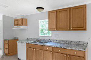 A kitchen with light wood cabinets, marble-patterned countertops, a double sink beneath a window, a dishwasher, and a range hood on the left wall. White walls and ceiling with a flush-mount light fixture are visible.