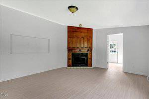 A bright living room with pale wood flooring, white walls, a wooden fireplace in the corner, and a ceiling light fixture. An open doorway leads to a sunlit room with a window.