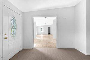 A view from the entryway of a house showing a white front door with a decorative glass window, leading to a bright, open room with tile flooring and white walls.