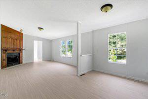 Bright living room with light-colored flooring, white walls, two windows, and a wooden fireplace on the left. Ceiling-mounted light fixtures are visible, and sunlight streams in through the windows.