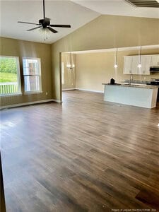 Spacious open-concept living area with dark wood flooring, beige walls, and vaulted ceiling. Large window on the left, kitchen with white cabinets and pendant lights is visible in the background.